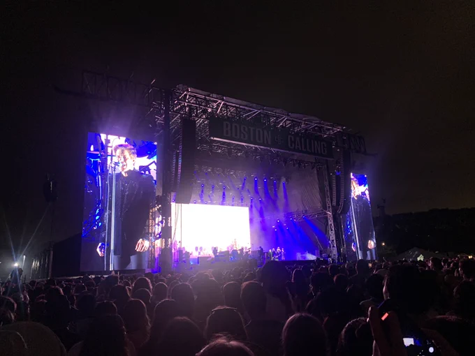 The Killers perform on the Boston Calling main stage. It’s late and dark under a warm evening sky. The lights are hazy. All you can see is the lead singer of the Killers on a big screen. He’s dressed in a black suit and standing singing. The rest of the stage is barely recognizable from this distance. It’s dark and bright.