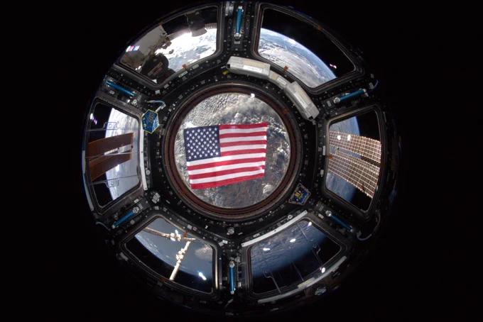 This image shows a fisheye view of the United States flag floating in the cupola of the International Space Station. Behind the flag, Earth is seen through the windows of the ISS. On the right and left side of the image, solar arrays are visible in space. Credit: NASA 