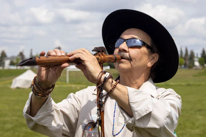 An Indigenous man wearing sunglasses, a wide-brimmed hat, a light shirt and both beaded and braided bracelets plays an ornate flute in Tawa Park.