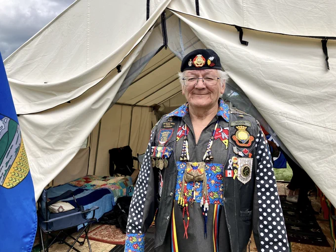 An older man with white hair, eyeglasses and a beret stands before the opening of a light-coloured tent pitched outdoors. He wears a dark shirt featuring floral and bison imagery, and a leather vest with several patches, pins and medals.