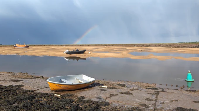 A picture of a coastal inlet dotted with small boats, with a dramatic sky overhead. Sunbeams are breaking through dark grey stormclouds, and a rainbow is touching the horizon. 