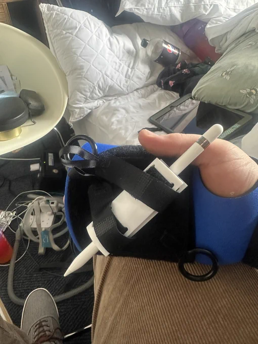 A white Apple Pencil held in a black and blue aid that is attached to my hand.