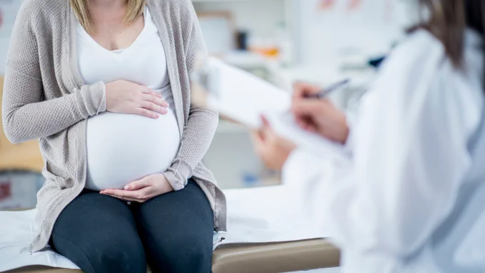 A shot of a pregnant women sitting on an examination table holding her belly with her right hand resting on top and her left below. Her head and lower legs are out of the frame. A doctor faces the patient and writes on a clipboard, while standing mostly out of the frame.