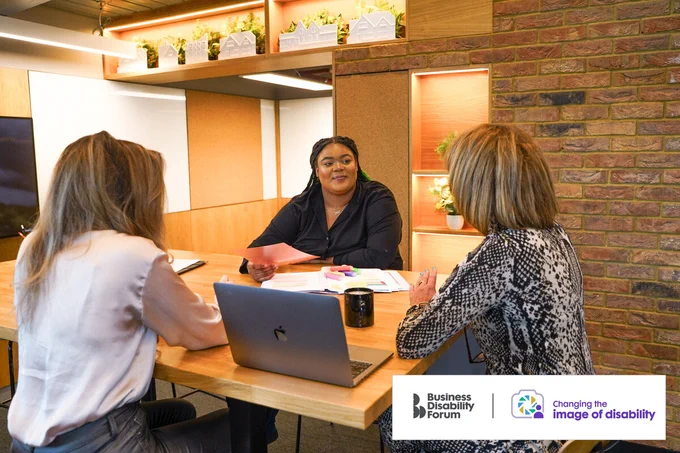 Professional with less-visible disability meets with 2 colleagues around a table. There are documents, devices and a coffee cup in front them.