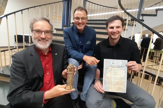 Drei Herren halten den Preis (eine Bronzestatue) und eine Urkunde in die Kamera.