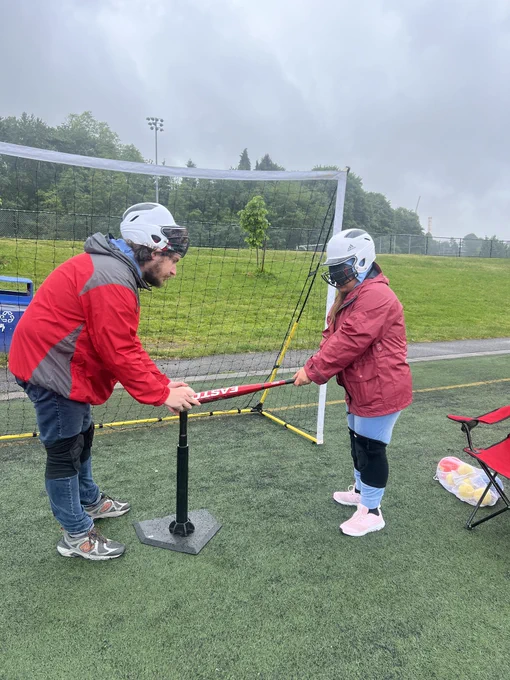 NWABA athlete is gripping a baseball bat at hip level as a volunteer holds onto the tip and aligns it with the top of the tee. Both people are wearing batting helmets and knee pads for safety.
