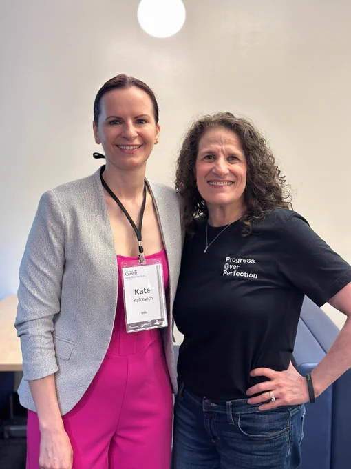 Kate in a pink jumpsuit and grey jacket with hair pulled back and me in a black progress over perfection shirt