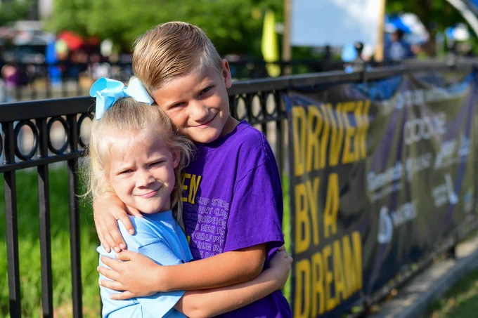 A light-skinned girl wearing a blue t-shirt and hair bow hugs a light-skinned boy wearing a purple "Driven by a Dream" t-shirt.