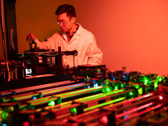 Daguan Nong (an East Asian man in a lab coat) is adjusting a microscope that has many metallic cylinders on a lab bench as part of its setup. 
