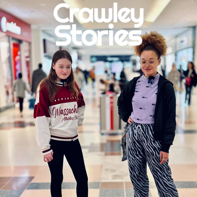 A photo of two people standing in a shopping centre, both facing the camera, their gazes angled downwards as they smile to themselves. At the top of the image is the white Crawley Stories logo.