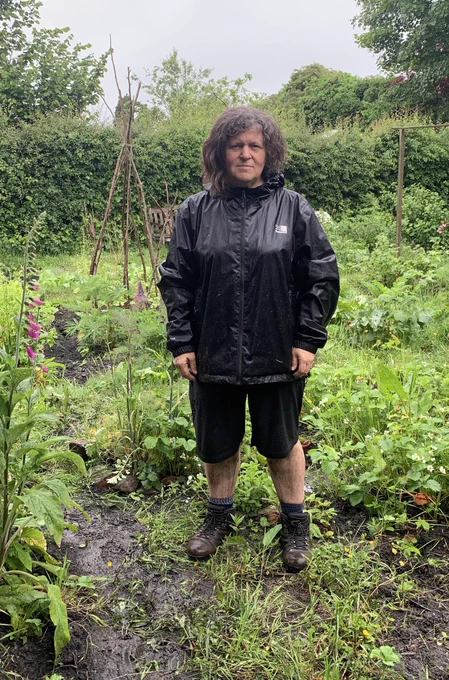 Photo of a person in black shorts & black waterproof coat wearing walking boots: the person looks very wet & muddy