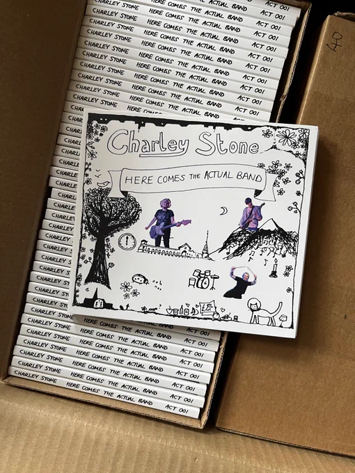 A CD of Here Comes The Actual Band by Charley Stone, on top of an open box of said CDs. Cover is a b&w line drawing with copy-pasted colour photo clippings of a guitarist, a drummer and a bassist, in a scene including a tree, a mountain and the Alexandra Palace.