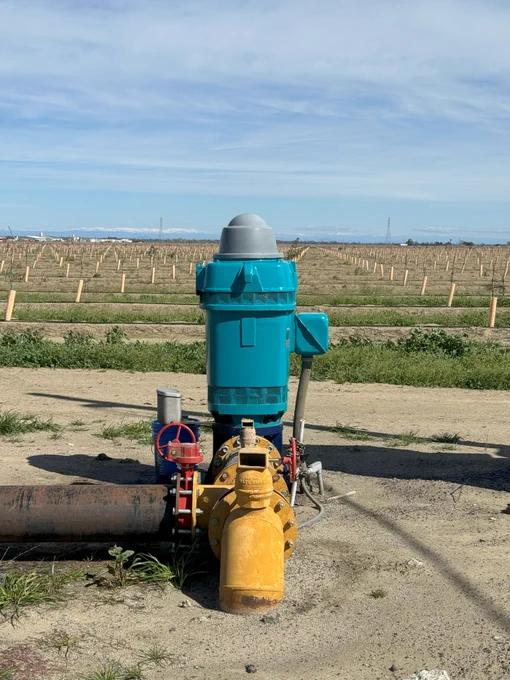 A blue and yellow groundwater well pump in foreground with a field of saplings planted in background.