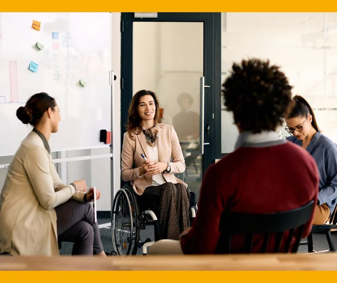 A group of people sitting in a meeting in an office. They are smiling and having a chat. The person facing the camera is a wheelchair user and leading the meeting.