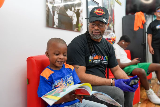 Student reads book with a barber at the NYC Reading Hub Ribbon Cutting at Nuevo Concepto Barber Shop on Harlem.