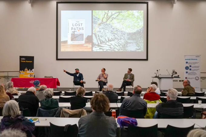 an audience facing a stage. I'm on the stage in a jacket and a hat pointing off to my right. Next to me are sitting Glen O'Hara and Tom Breen from the In all our footsteps project. 
