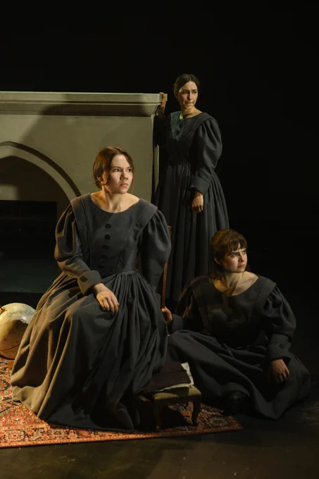 Three actors in period dress sitting on a rug and looking camera with tense looks on their faces. 