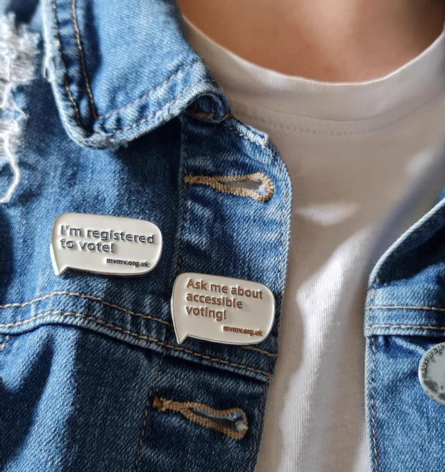 Two enamel pin badges on a blue denim jacket. Both are shaped like speech bubbles. One says I'm registered to vote mvmv.org.uk in navy blue text. The other says Ask me about accessible voting mvmv.org.uk in orange text.