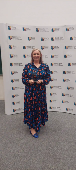 A woman (Mags) is standing in front a a Maynooth University background. She is holding her trophy and smiling at the camera.