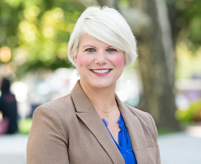 Professional portrait of a smiling person with short blonde hair, wearing a tan blazer over a blue blouse. The background features a softly focused park setting with trees and a walking path.