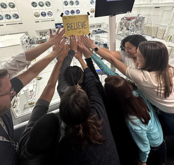 Several members of the Europa Clipper team stand below a yellow Ted Lasso "Believe" sign that has been taped to the window of the gallery overlooking the Europa Clipper clean room at NASA JPL. They are leaning into the sign and have all placed their hands on it.