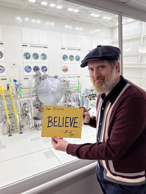 Brendan Hunt, also known as "Coach Beard" on Ted Lasso, stands in the gallery overlooking the Europa Clipper clean room at NASA JPL. He is holding a yellow "Believe" sign that he has autographed for the Europa Clipper team.
