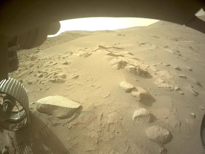 A beige-colored image shows the rocky and sandy Martian surface underneath the Perseverance rover. On the far left is its back wheel and it shows a small rocks inside of it.