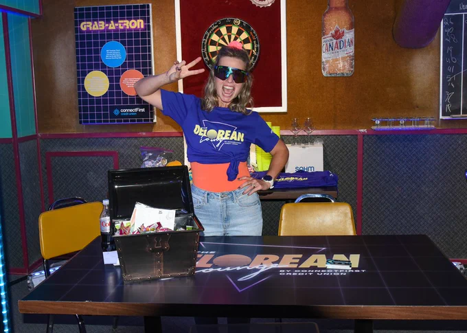 A connectFirst staff member is posing for the camera in front of a dart board at the #1 Royal Canadian Legion. There is a table in front of her that reads "DeLorean Lounge by connectFirst Credit Union"
