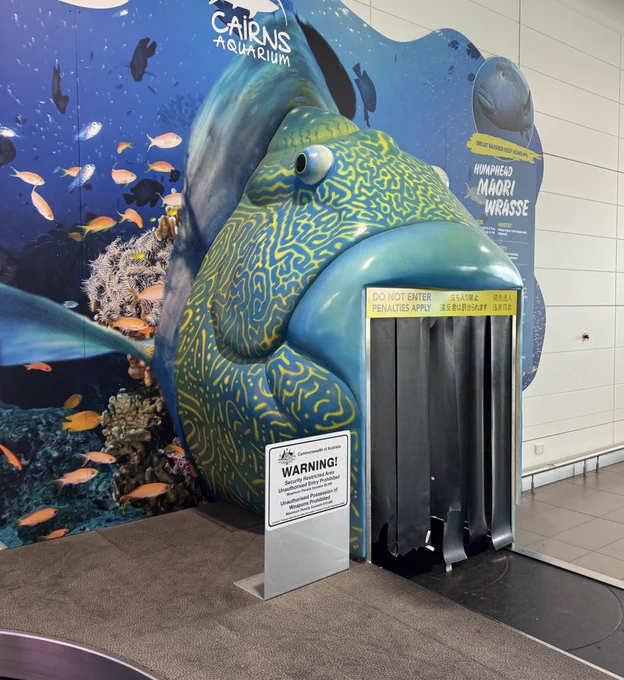 A large fish at baggage claim is posed to barf bags onto the belt. It’s large and blue with yellow twisty patterns