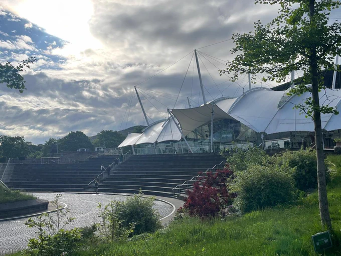 Our Dynamic Earth science center in Edinburgh, featuring a futuristic, tent-like structure with multiple conical roofs supported by cables. Surrounding the main building are lush green trees, a circular patterned pavement, and a series of steps leading up to the entrance, all under a partly cloudy sky.