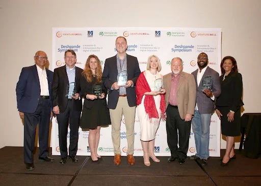 Group of eight individuals holding awards at the Deshpande Symposium, standing in front of a banner with logos including VentureWell and UMass Lowell.