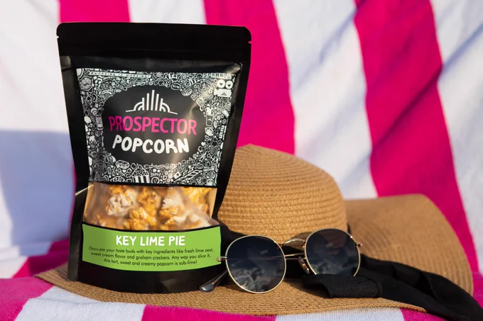 A bag of Key Lime Pie gourmet popcorn sits next to a sun hat and glasses on a beach towel with pink and white stripes.