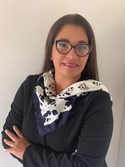 A headshot of Maria. She is wearing a dark long-sleeve shirt, a blue and white silk scarf, and glasses. 