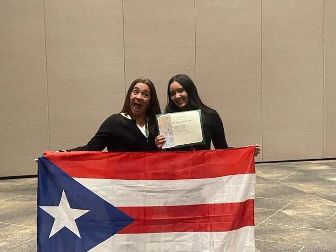 A photo of Maria and a student holding up the flag of Puerto Rico and an award certificate. 