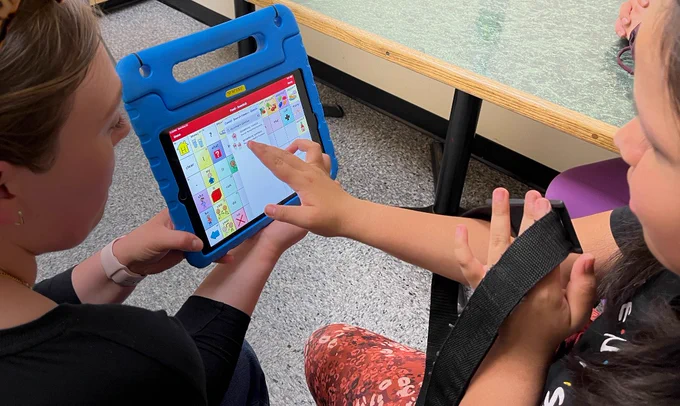 a woman holds an iPad with TouchChat edit screen visible.  The student points finger at screen to select.