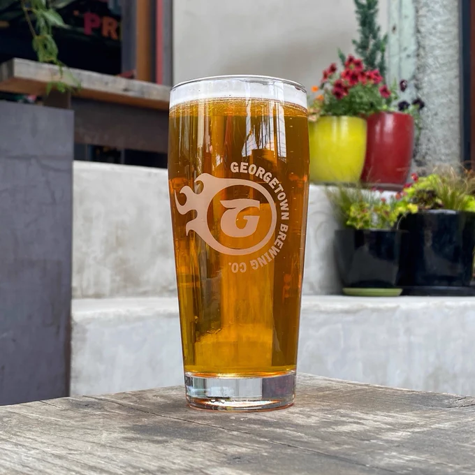 A pint glass of a clear golden beer with a foamy head sits on a wooden table in the beer garden.