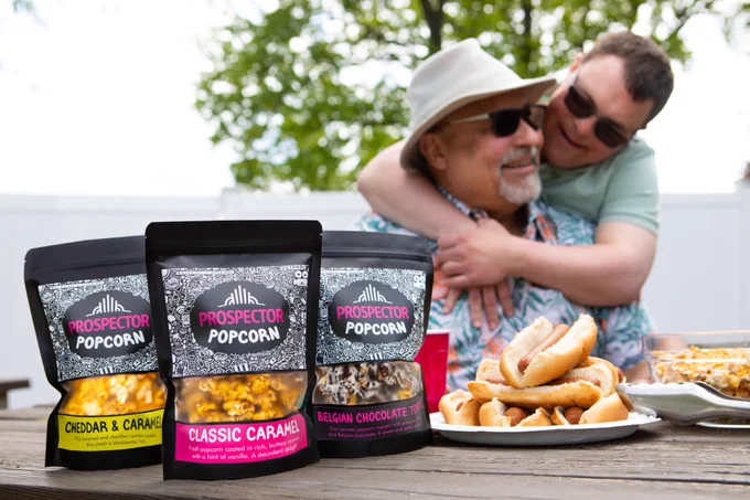 A father and son hug at a barbecue. Hot dogs and gourmet popcorn can be see in the foreground.