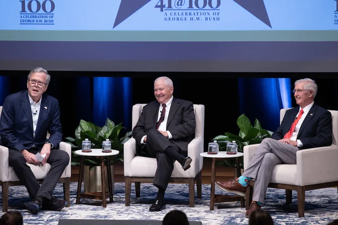 Jeb Bush, Robert Gates, and Andy Card are seated on stage at an event titled "A Celebration of George H.W. Bush," commemorating the 100th anniversary of George H.W. Bush's birth. They are in a conference setting, each seated on individual armchairs, with a backdrop displaying the event's title and the seal of the President of the United States.