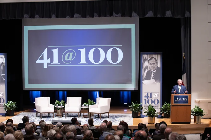 President Mark Welsh speaks at a podium during an event commemorating the 41st President, represented by the theme "41 at 100," in a packed auditorium with a large screen displaying the event's logo, and banners featuring the same logo flanking the stage.