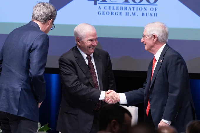 Robert Gates and Andy Card shaking hands on a stage at an event celebrating George H.W. Bush, with Jeb Bush standing nearby observing the handshake. The background features a banner with the text "A Celebration of George H.W. Bush.