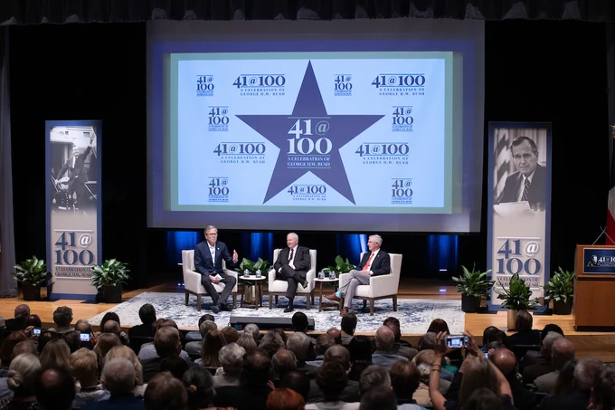Jeb Bush, Robert Gates, and Andy Card participate in a panel discussion in a crowded auditorium, seated in front of a large screen displaying "41@100" logos commemorating the 100th birthday of George H.W. Bush, with his portrait visible.