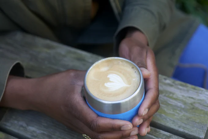 A photo looking down on someone holding a resuable mug with both hands. The mug is filled with a coffee that has a foam heart decorated on top.