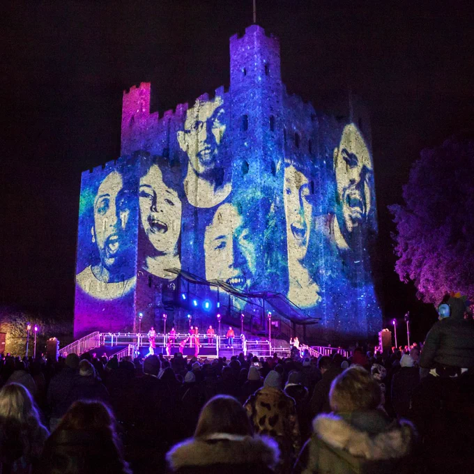 Large faces projected onto the side of Rochester Castle.  A crowd of people stands in front looking up at the projected image.
