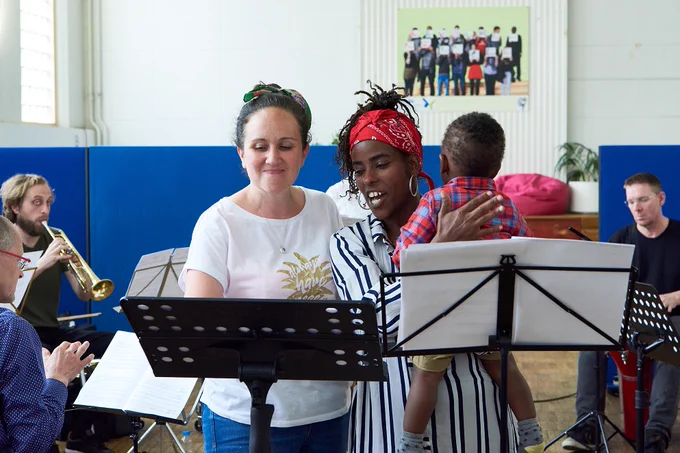 A photo of two women in front of music stands, one holding a baby; musicians in the background including a trumpet player - photo by Micha Theiner