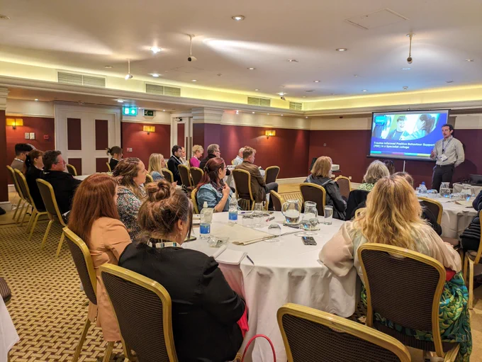 A room full of people listening to a white man present at the front of the room.