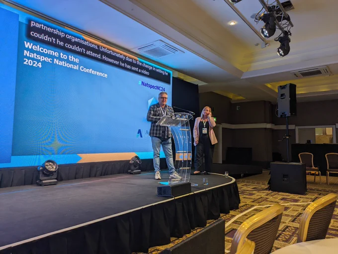 Two people, one a middle aged white man and the other a similarly aged woman, standing on the Natspec stage to present.