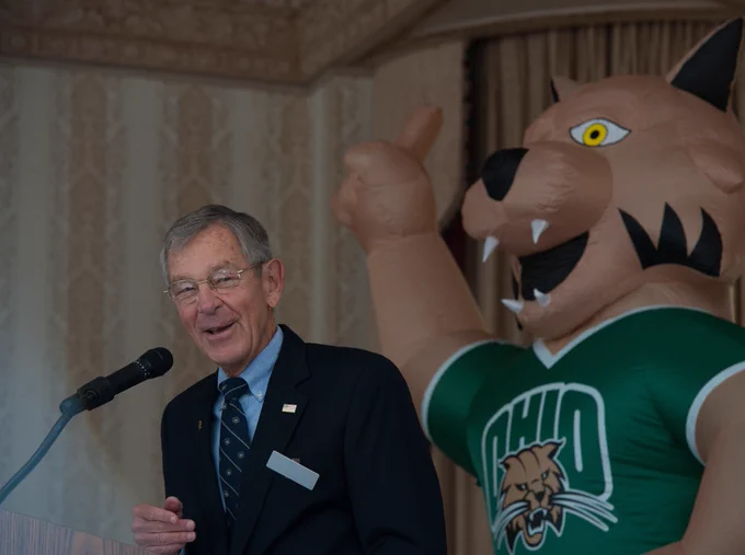 A man in a suit stands at a podium speaking, with a large inflatable mascot of the Ohio Bobcats behind him, gesturing with a raised arm.