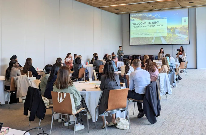 A group of participants sitting down at tables and viewing a PowerPoint presentation at the front of the room. 