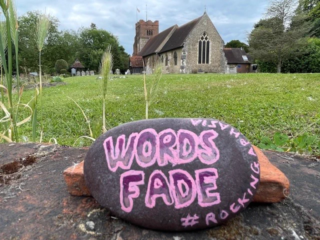 Picture of a painted rock with the hashtag '#RockingAphasia'. The rock is in front of a Norman church. 