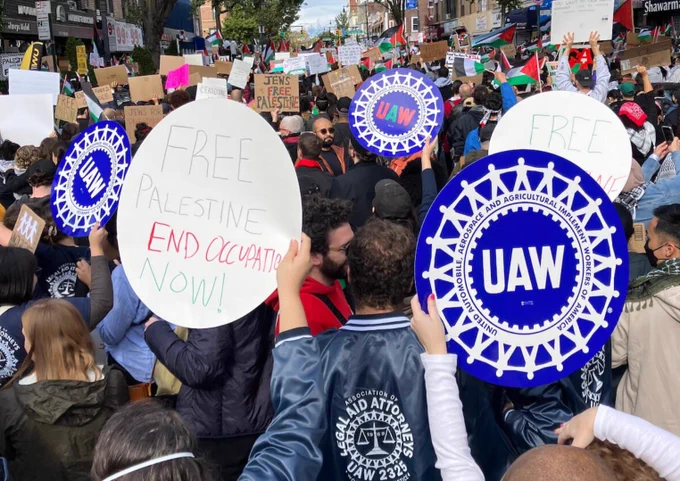 Photograph of a protest. In the foreground is a white circular sign which reads, “Free Palestine, end occupation now!” On the right is a circular blue sign with the UAW logo on it.